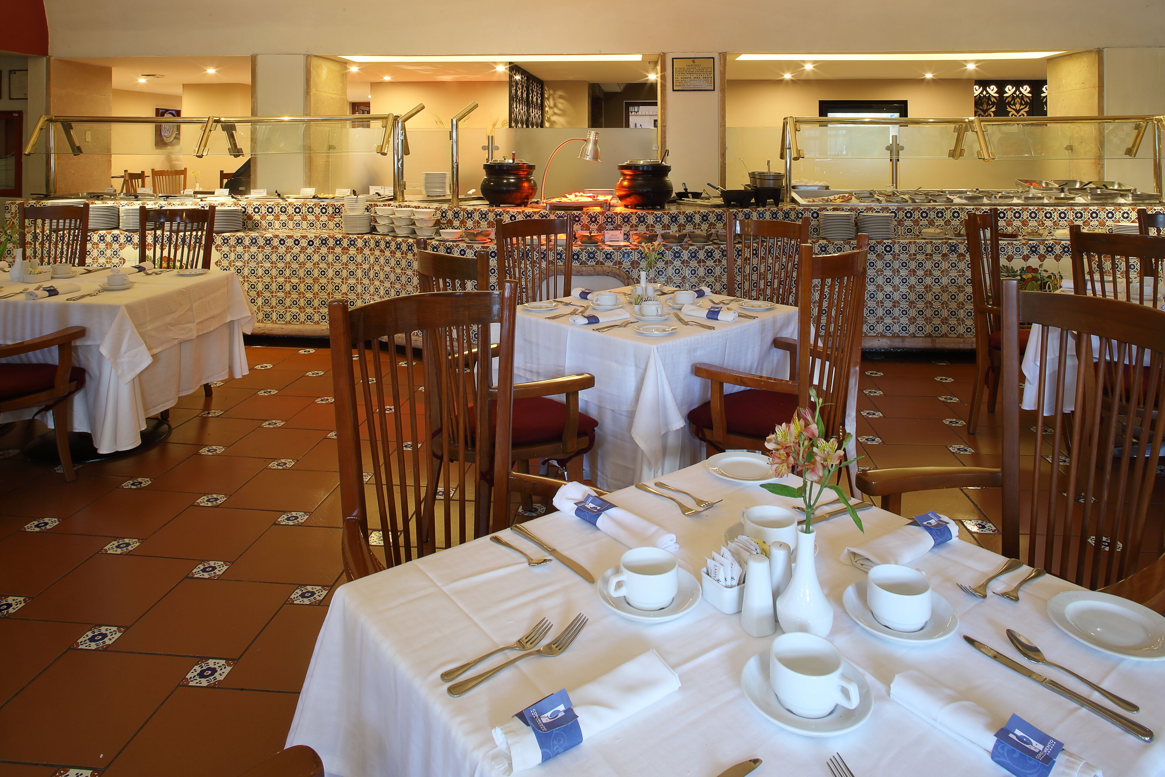 A dining area with tables set for breakfast, featuring white tablecloths, wooden chairs, and neatly arranged breakfast items.