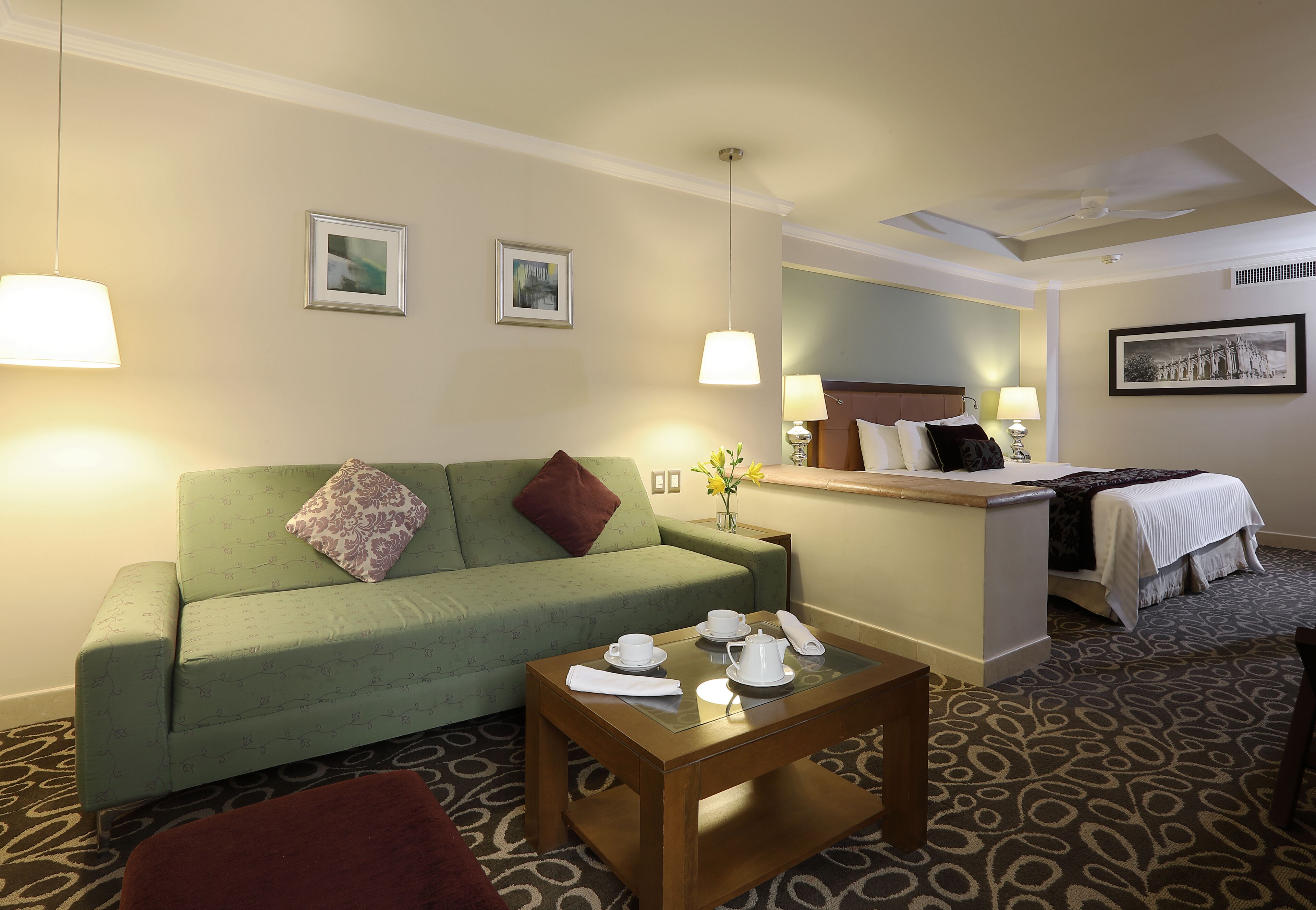 Habitación de hotel con un sofá verde, una mesa de centro de madera con juego de té, una cama con edredón blanco y una mesita con un jarrón de flores.