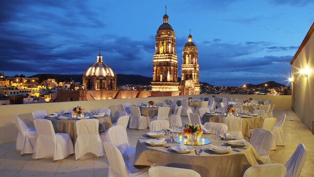 Un área de comedor al aire libre con mesas dispuestas para un evento formal, con vistas a un edificio histórico con dos torres abovedadas.