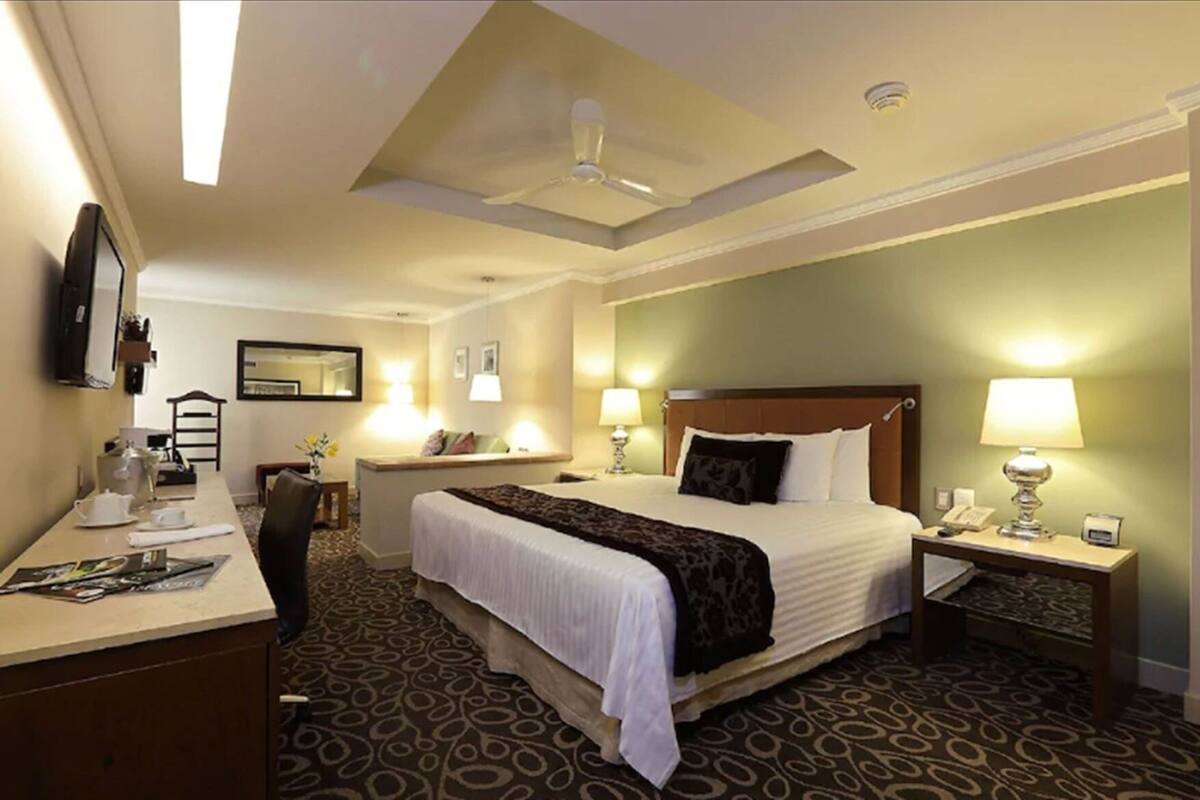Habitación de hotel con cama, mesitas de noche, escritorio con silla, ventilador de techo y ventana grande.