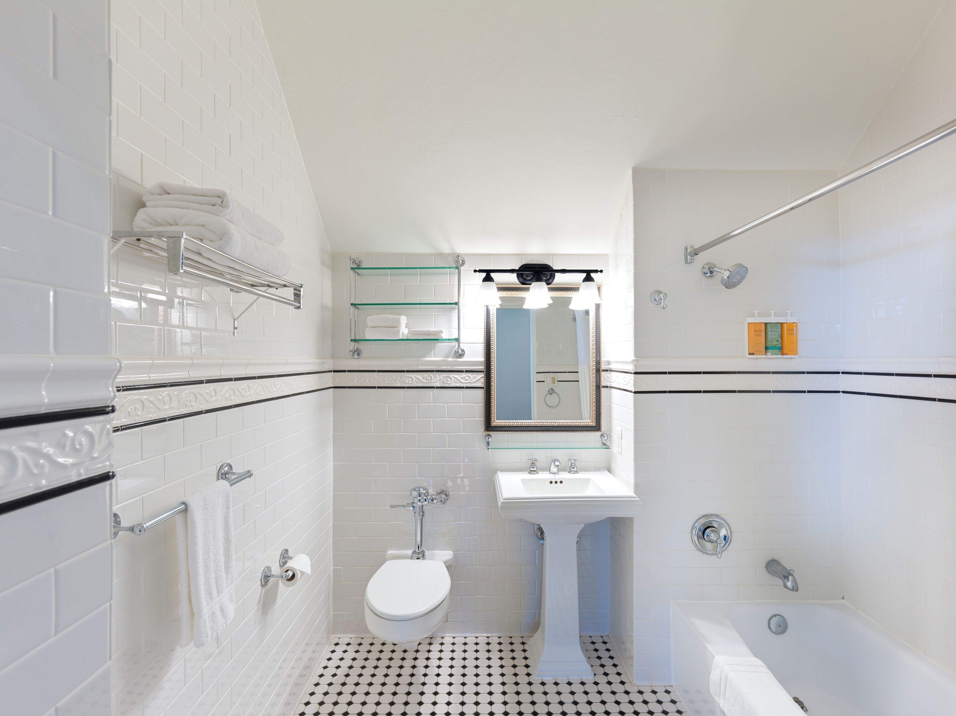 Un baño con inodoro, lavabo y bañera. Hay un estante de vidrio sobre el lavabo y un perchero en la pared.