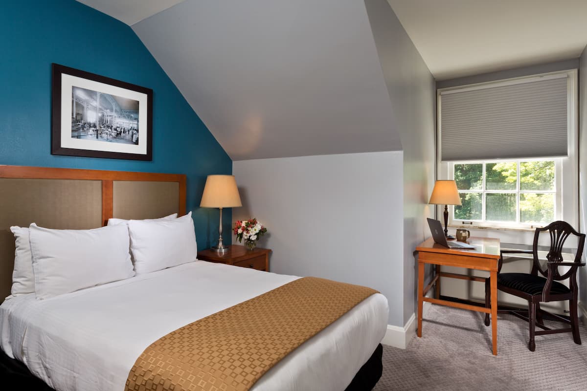 Une chambre d’hôtel avec un lit, un bureau, une chaise, une fenêtre avec des stores et un tableau encadré au mur.