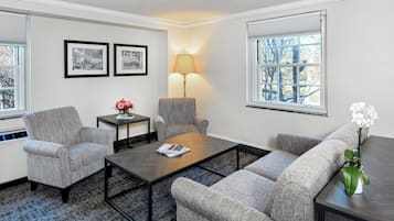 A modern living room with a sofa, armchairs, a coffee table, and a lamp. There are framed pictures on the wall and a window with blinds.