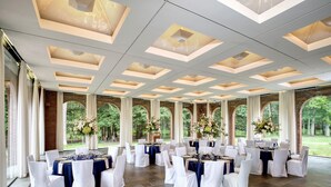 Une salle de banquet où les tables sont dressées pour un événement formel, avec un sol carrelé en pierre et de grandes fenêtres offrant une vue sur la verdure.