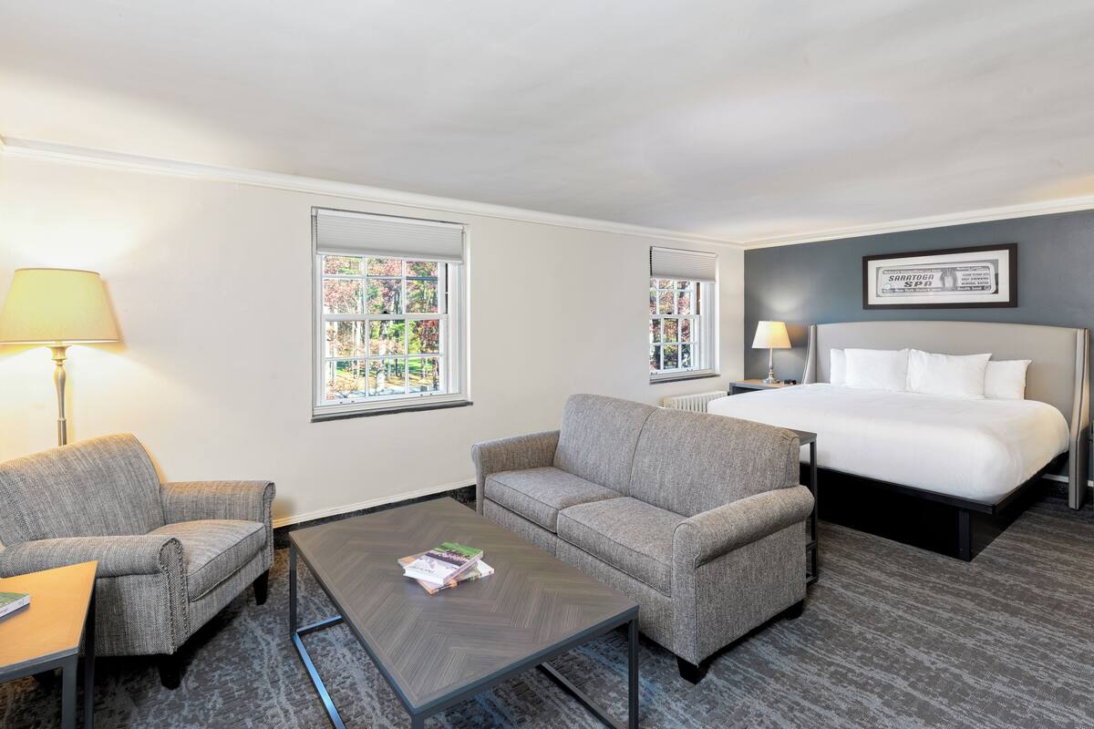 Une chambre d’hôtel comprenant un lit, deux fauteuils, une table basse et deux fenêtres équipées de stores.