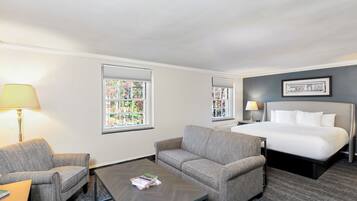 A hotel room with a bed, two armchairs, a coffee table, and two windows with blinds.