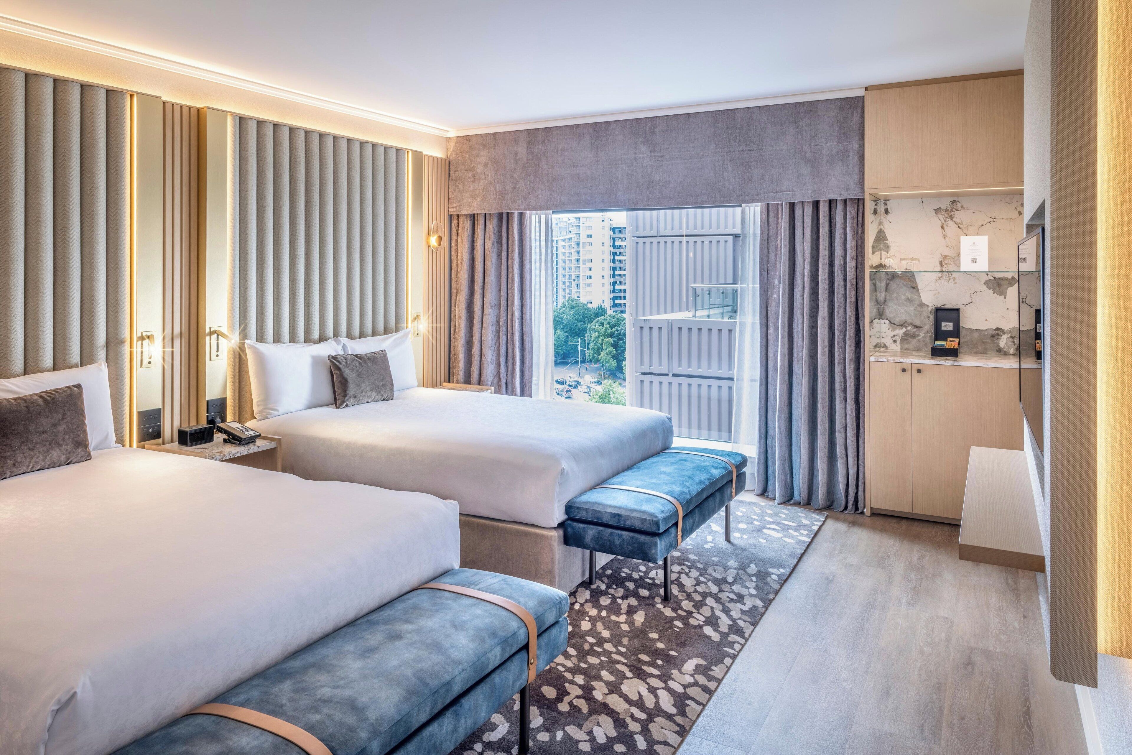 Habitación de hotel con dos camas, un banco, un escritorio y vistas a la ciudad.