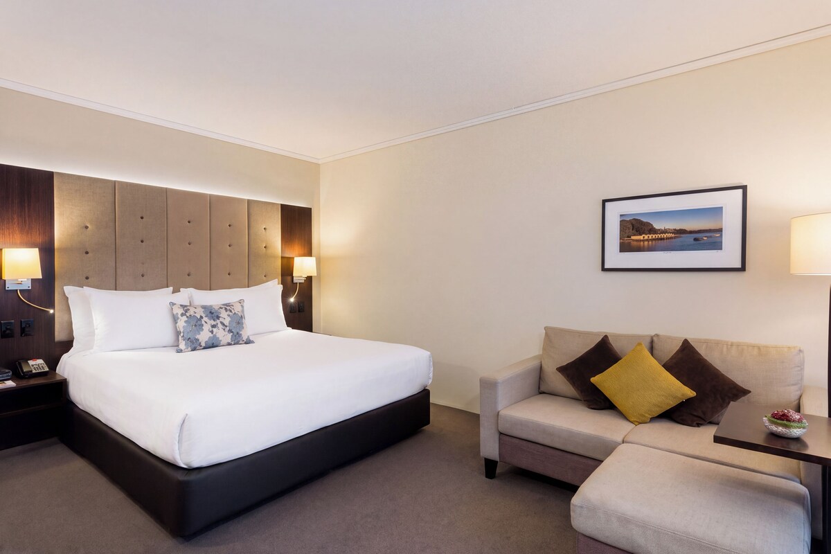A hotel room with a large bed, a sofa, a small table with a lamp, and a framed picture on the wall.