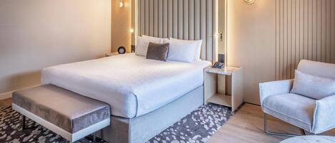 A modern hotel room with a large bed, bedside table, chair, and a small round table.