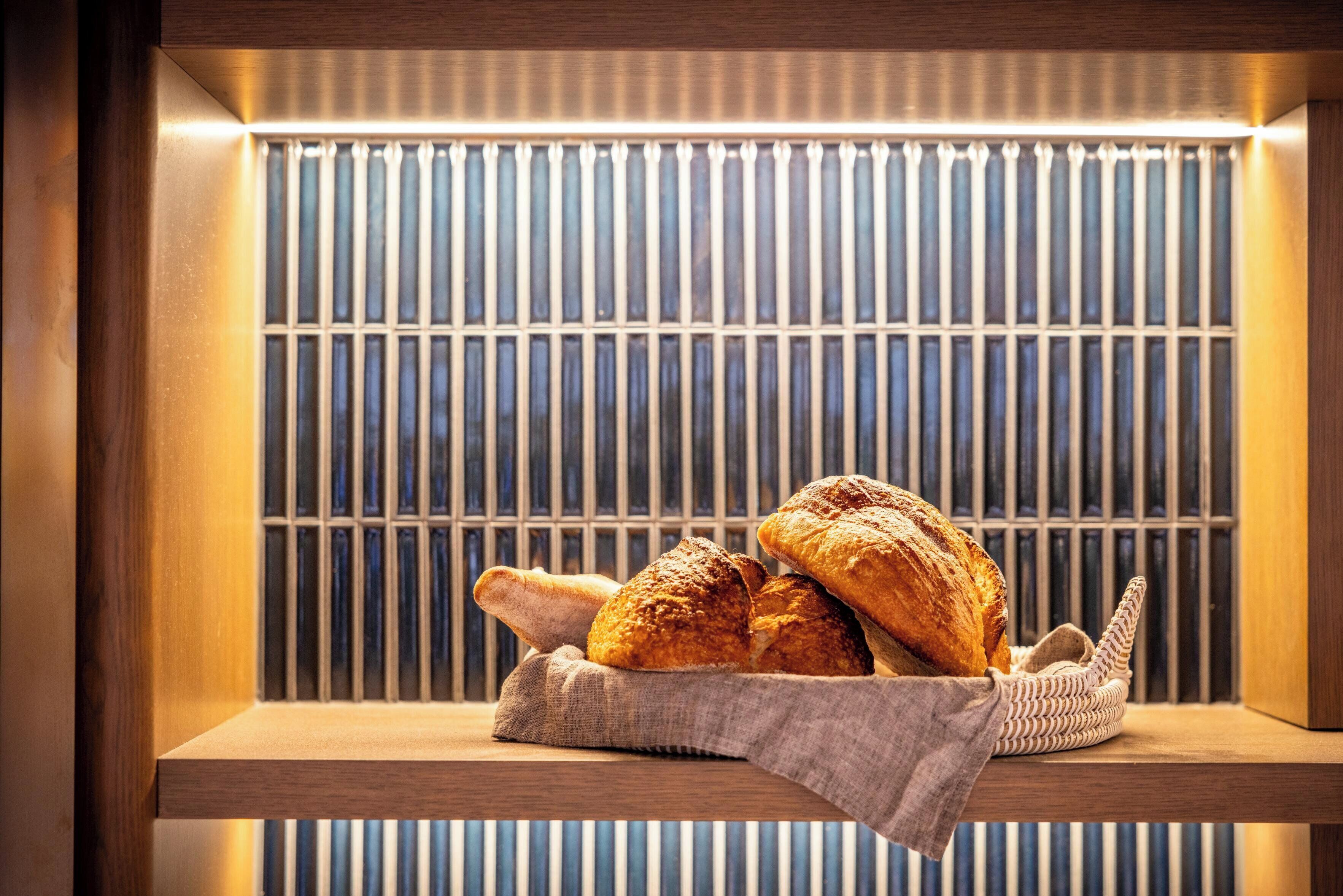 A wooden shelf with a basket containing two loaves of bread and a baguette, set against a background of vertical blinds.