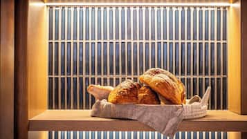 A wooden shelf with a basket containing two loaves of bread and a baguette, set against a background of vertical blinds.