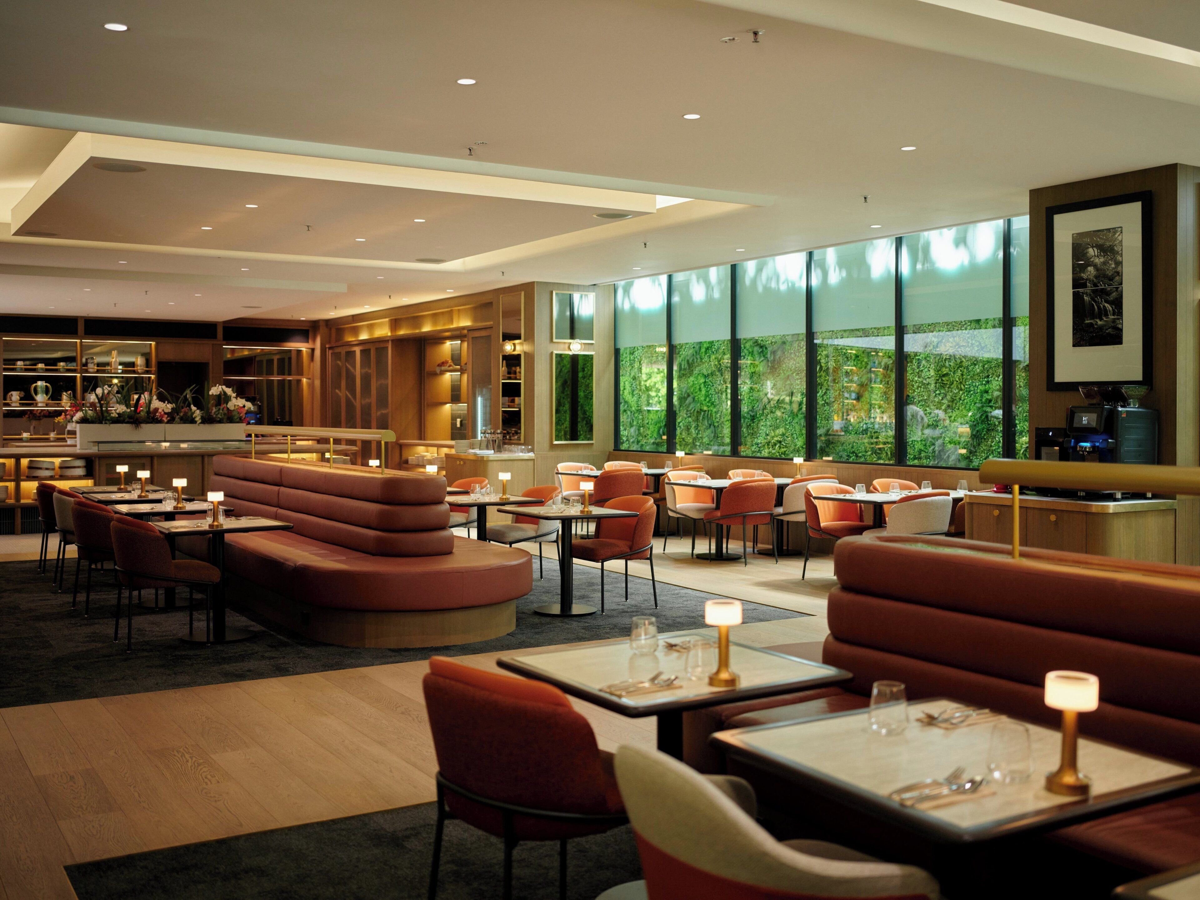 Un restaurant moderne avec des banquettes et des tables, un coin bar et de grandes fenêtres donnant sur un espace verdoyant.