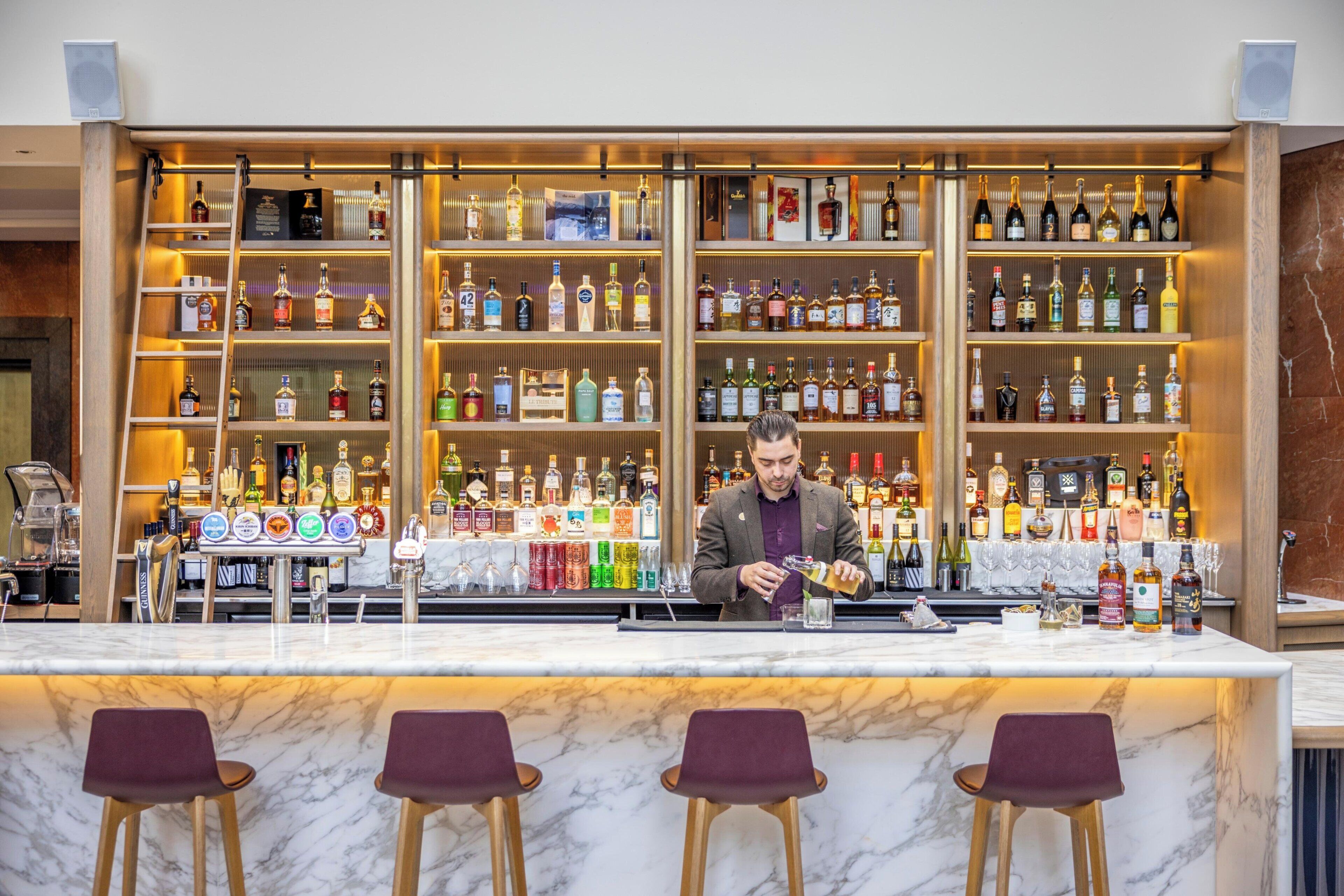 Eine Bar mit einem Barkeeper, der ein Getränk zubereitet, Regalen voller Spirituosenflaschen und Marmor-Barhocker.