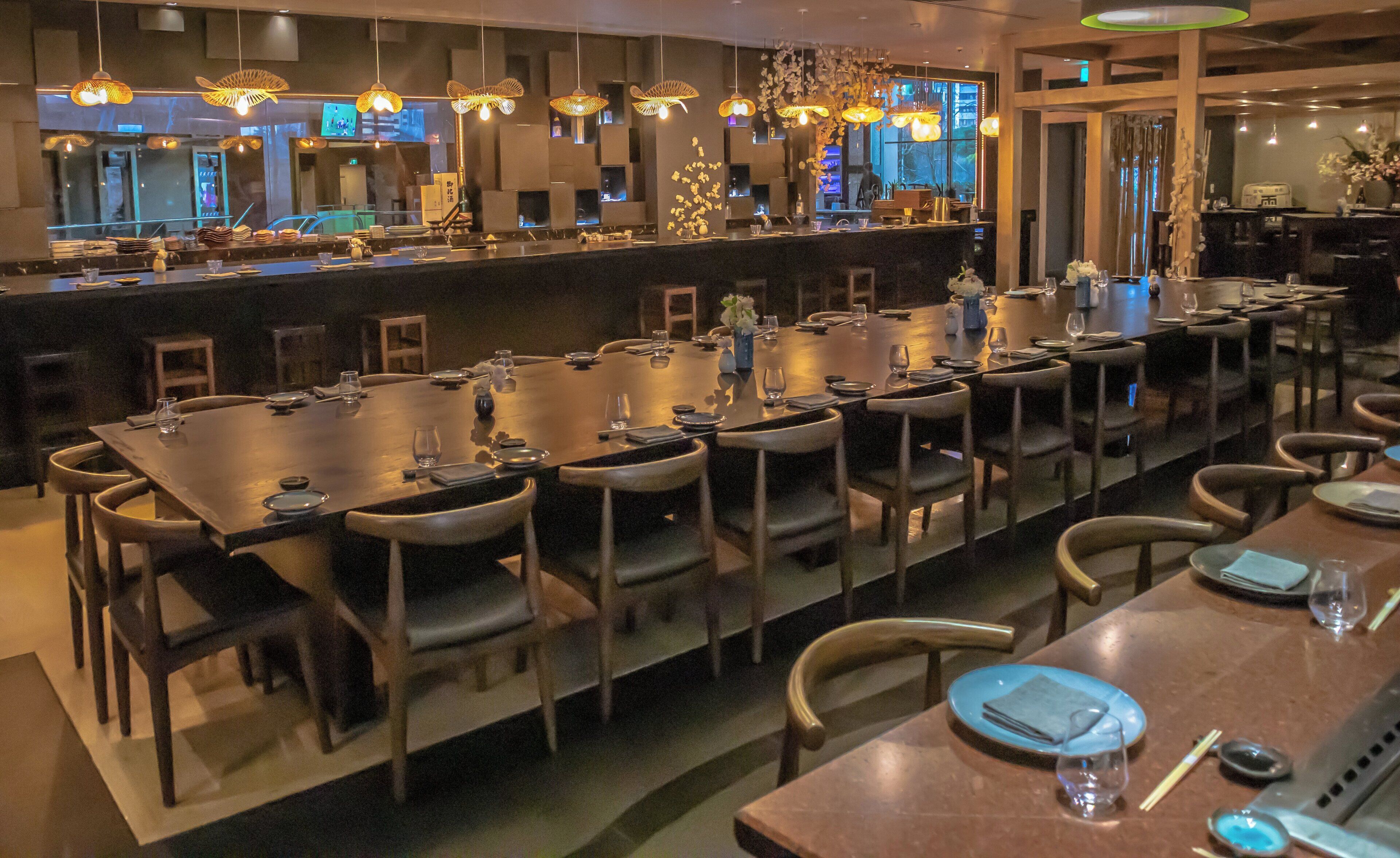 A restaurant with long communal tables, chairs, and hanging lights.