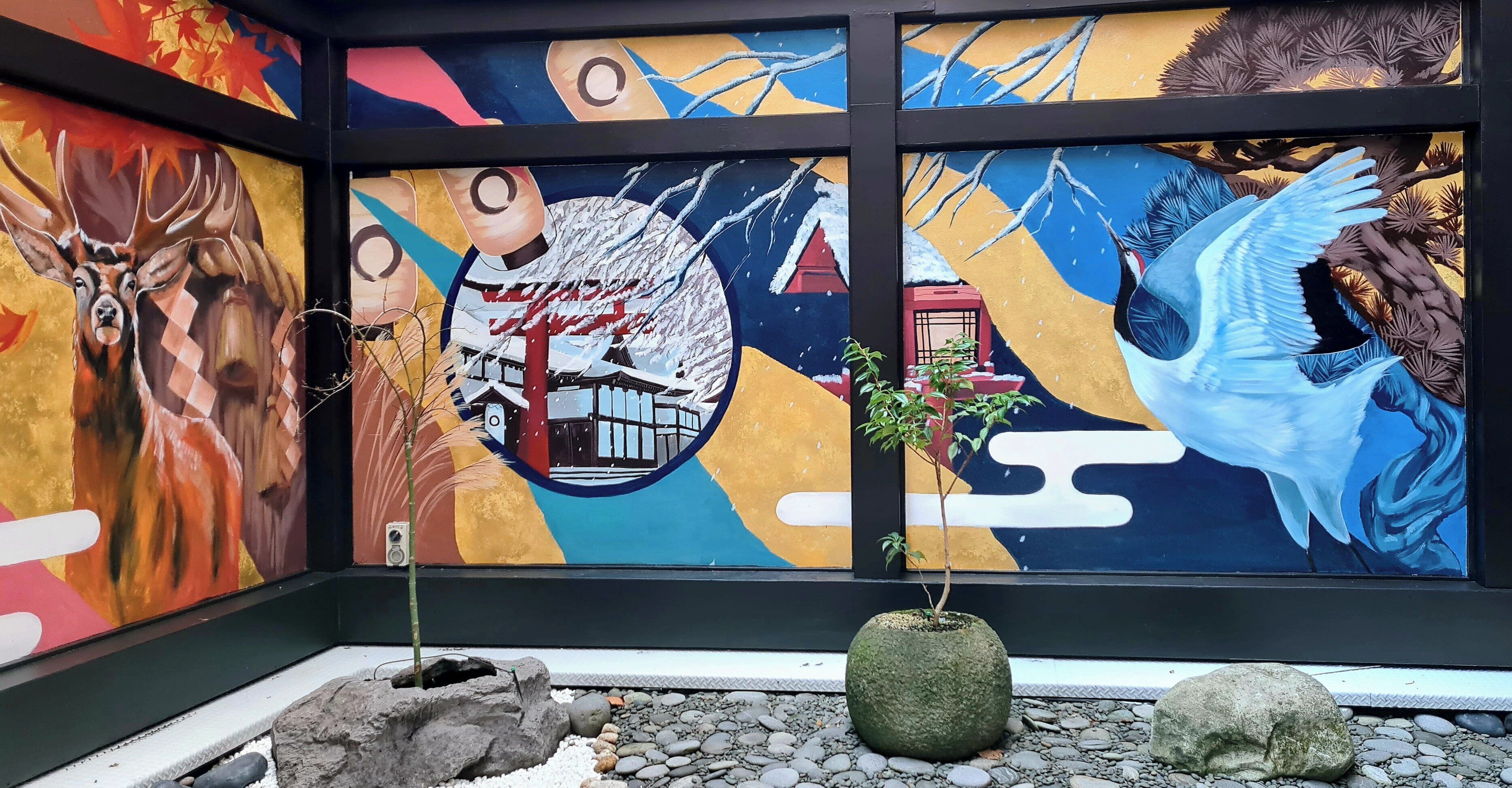 A mural with a deer, a bird, and a traditional building. There are two potted plants and a rock garden in front.