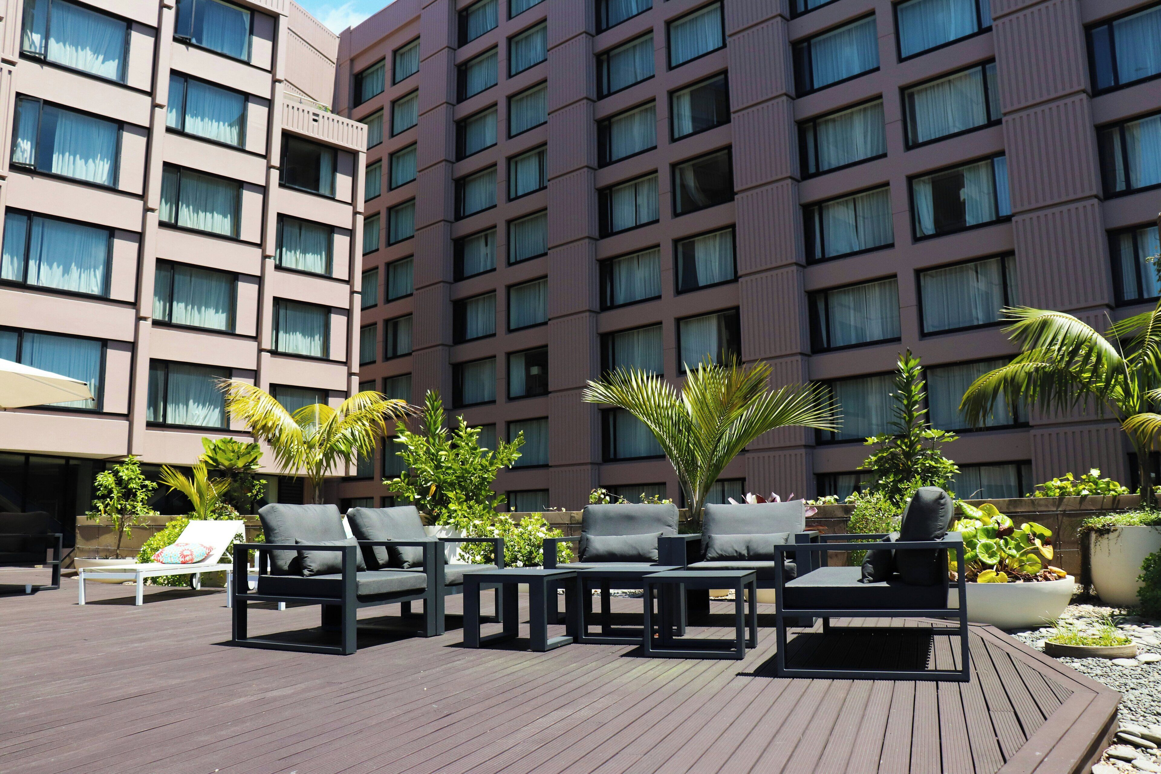 A rooftop patio with outdoor seating, including lounge chairs and tables, surrounded by potted plants and palm trees.