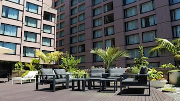 A rooftop patio with outdoor seating, including lounge chairs and tables, surrounded by potted plants and palm trees.