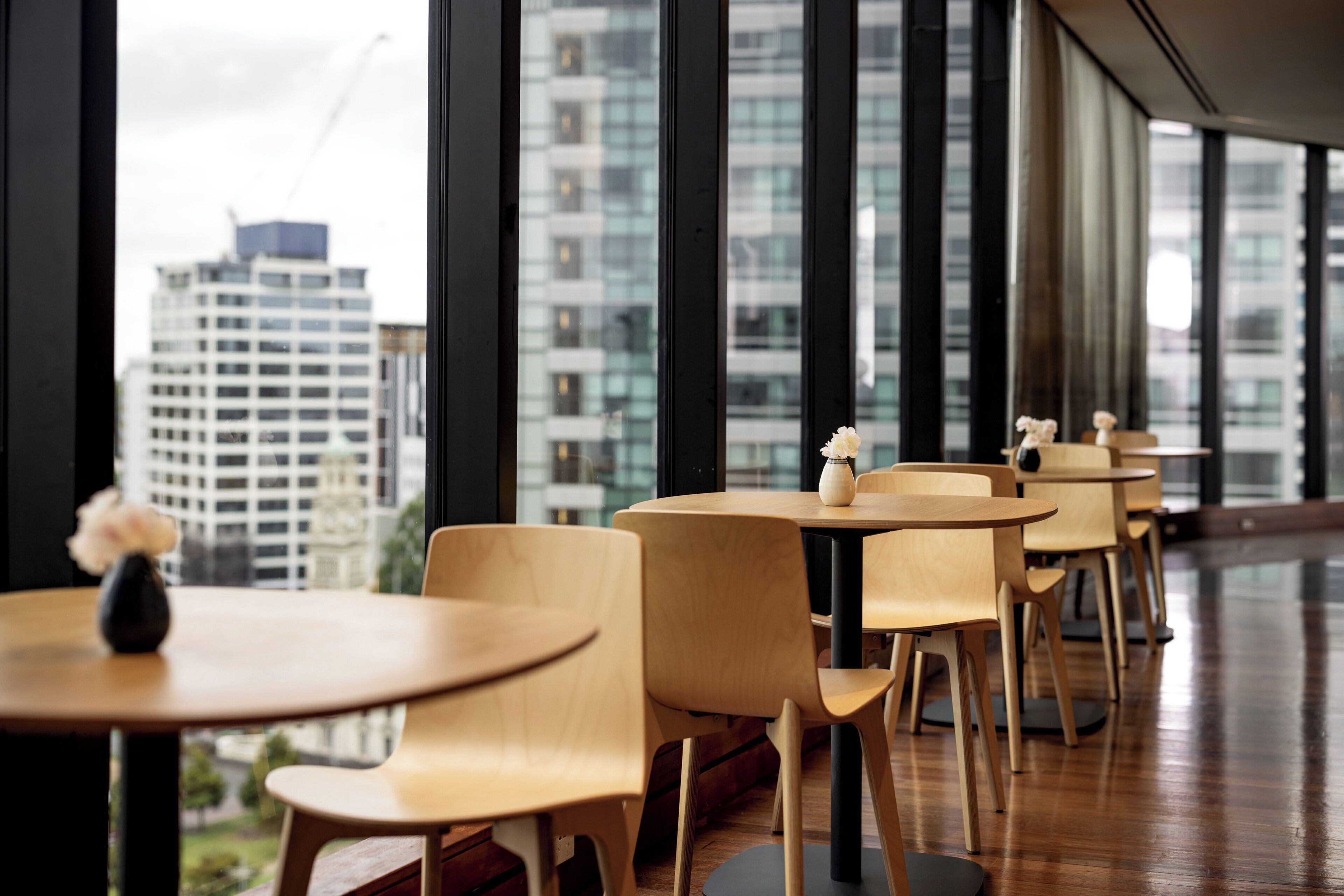 A modern indoor space with round tables and chairs, large windows offering a view of tall buildings.