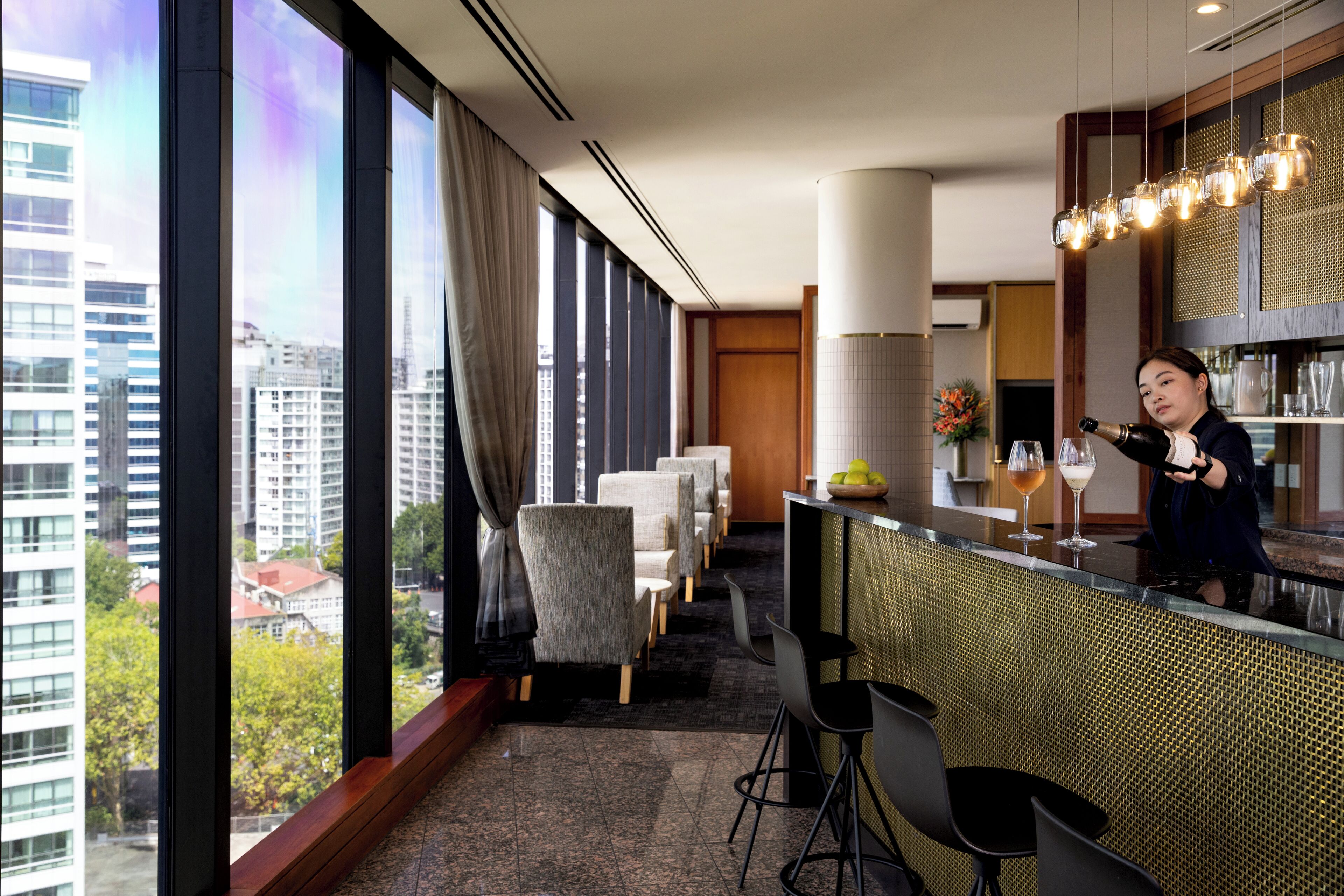 A modern hotel bar with a city view, bar stools, and a bartender.