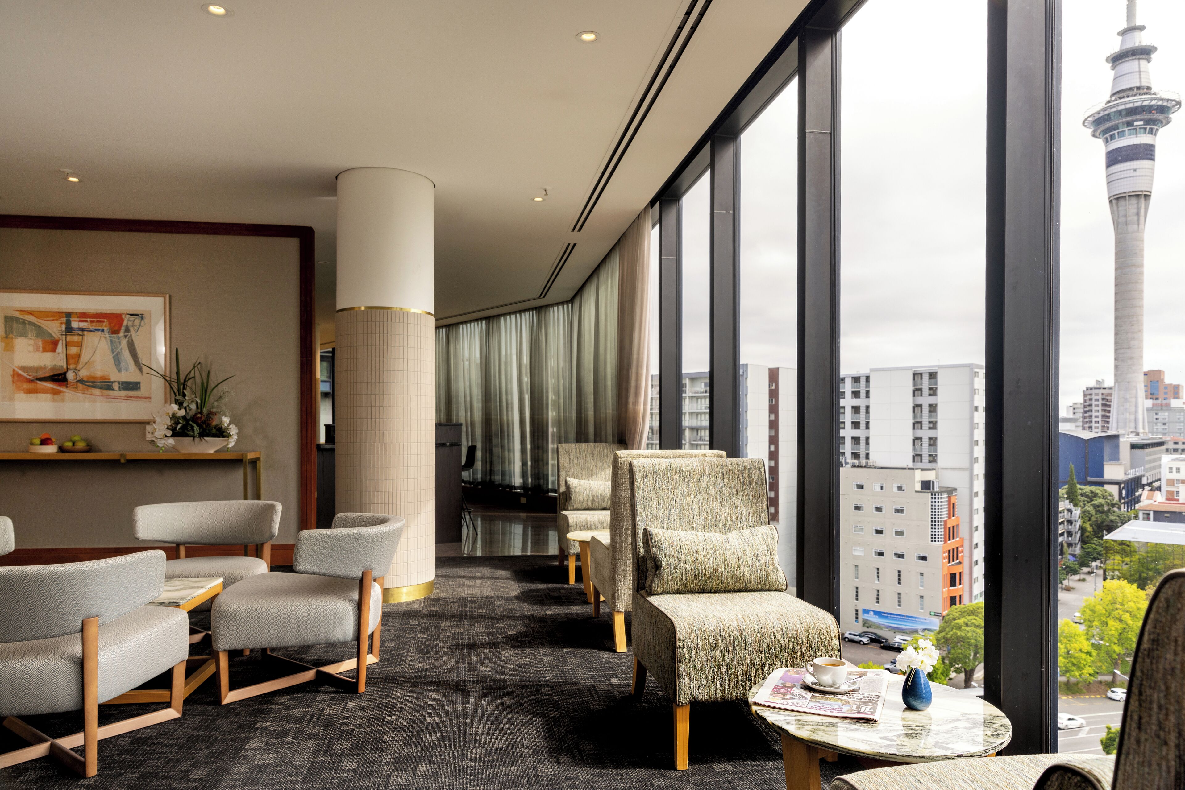 Vestíbulo de hotel con asientos, un mostrador de recepción y vistas a un paisaje urbano que incluye una torre.