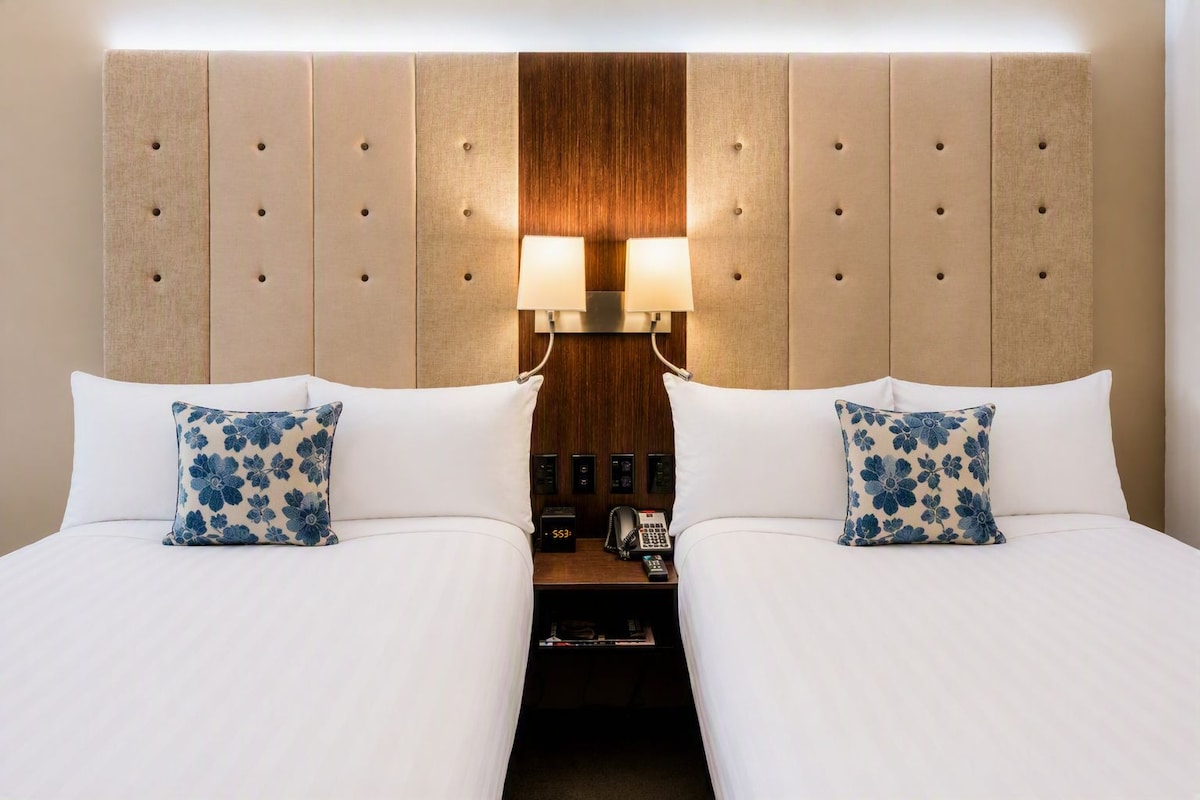 A hotel room with two beds, each with a white bedspread and a blue floral pillow. There is a wooden headboard with a light fixture in the center.
