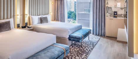 A modern hotel room with two beds, a bench, a small desk, and a view of the city.