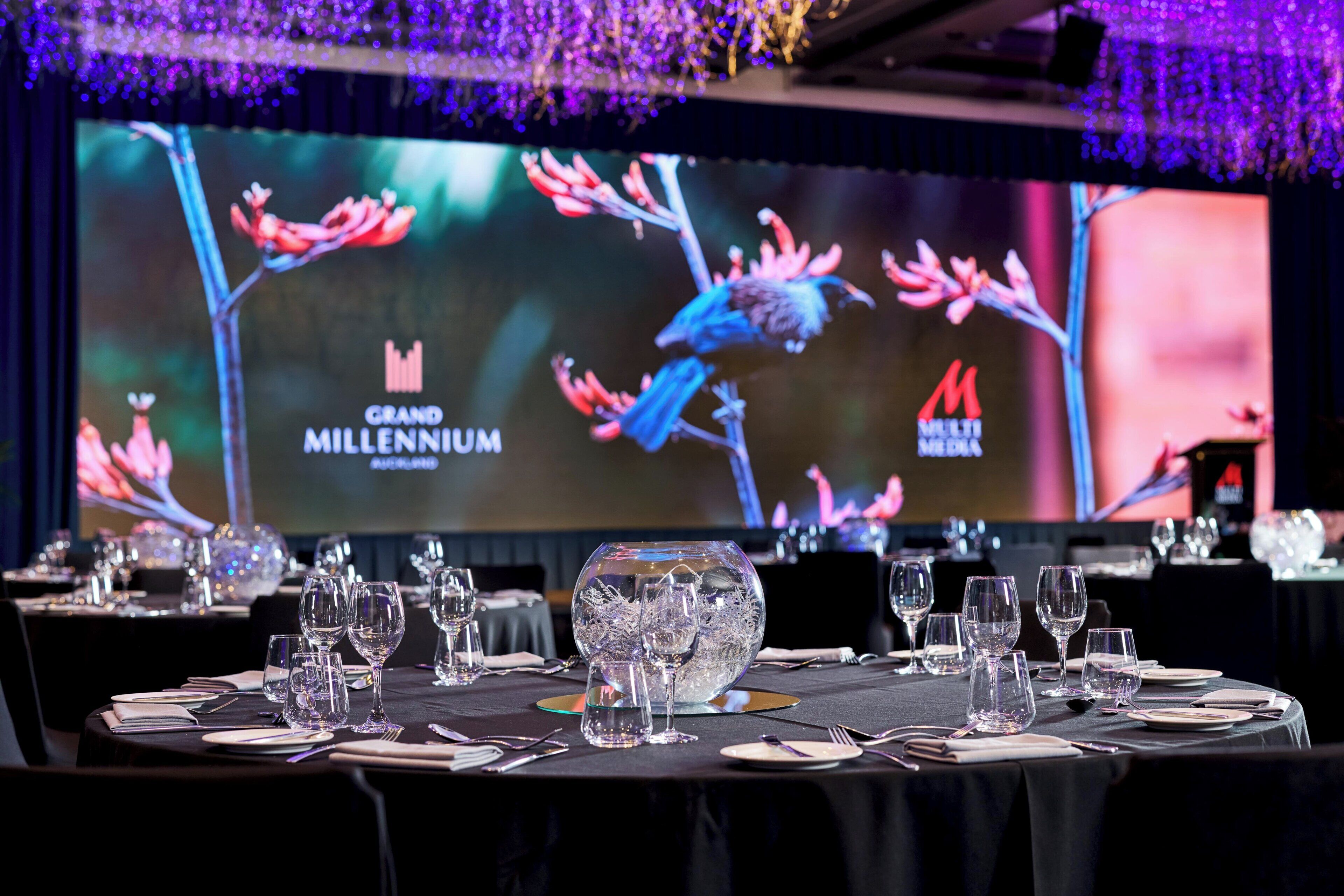Une salle de banquet où les tables sont dressées pour un événement officiel, avec un grand écran affichant les logos « Grand Millennium » et « MILLENNIUM ».