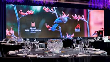 Uma sala de banquetes com mesas montadas para um evento formal, exibindo uma grande tela com os logotipos 'Grand Millennium' e 'MILLENNIUM'.