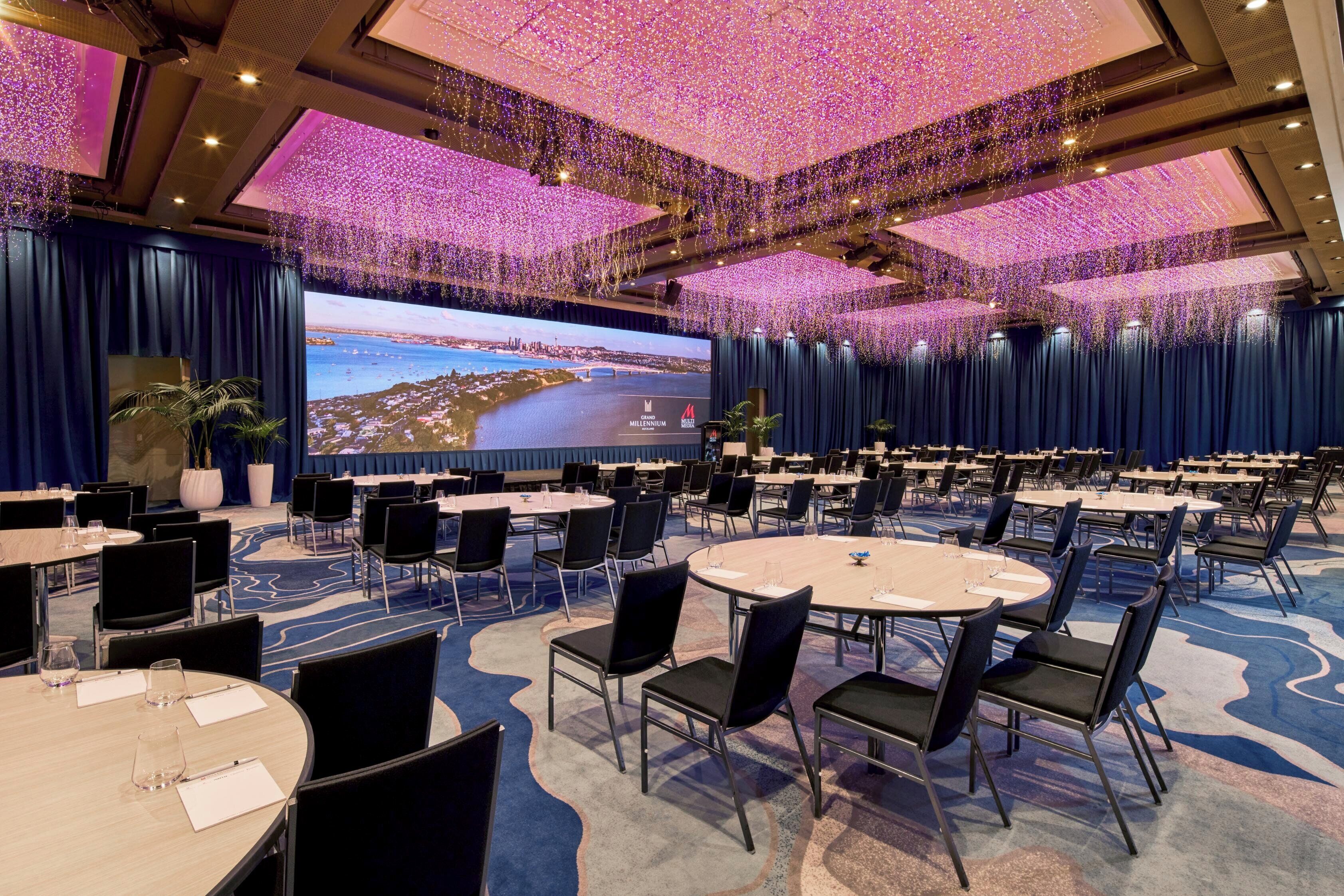 A conference room with round tables, chairs, a large screen, and a ceiling adorned with hanging lights.