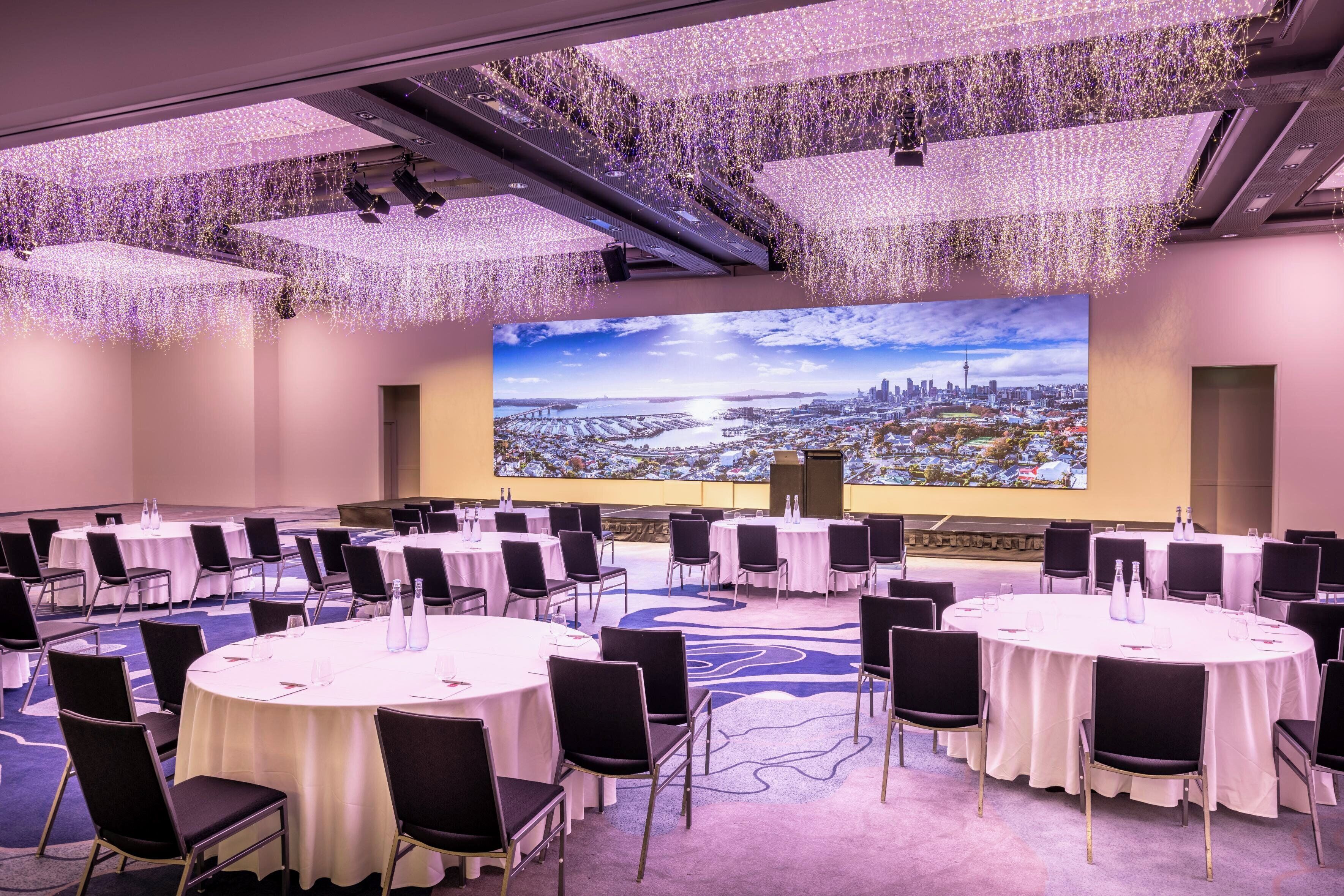 A conference room with multiple round tables, black chairs, and a large screen displaying a cityscape.