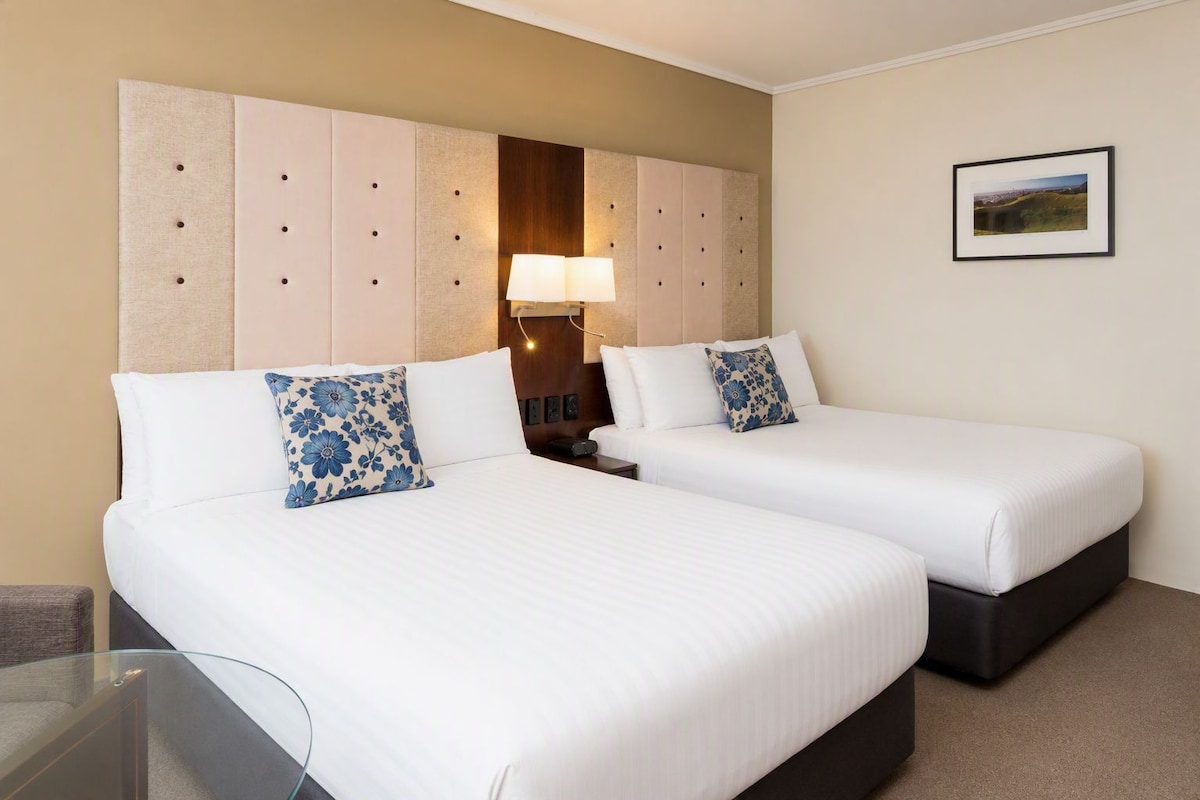 A hotel room with two beds, a large padded headboard, a bedside table with a lamp, and a framed picture on the wall.