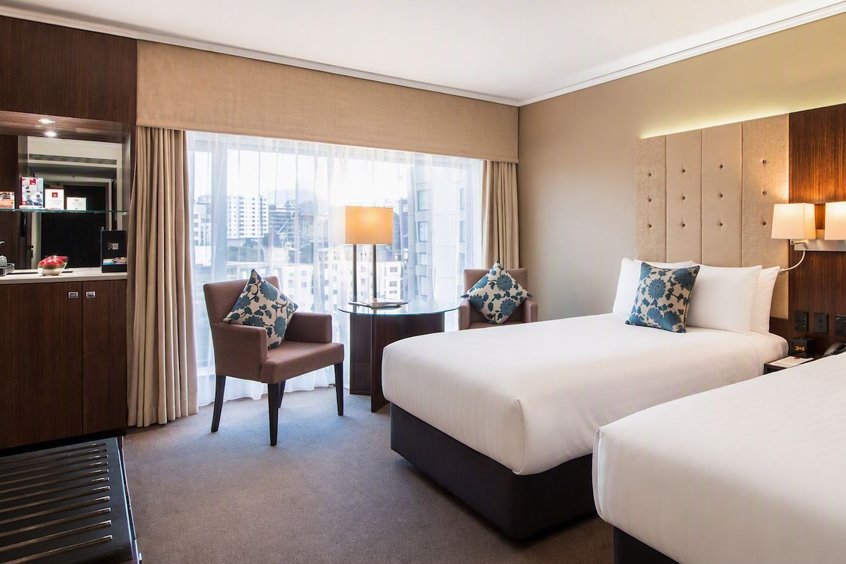 A hotel room with two beds, a desk, a chair, and a view of the city.