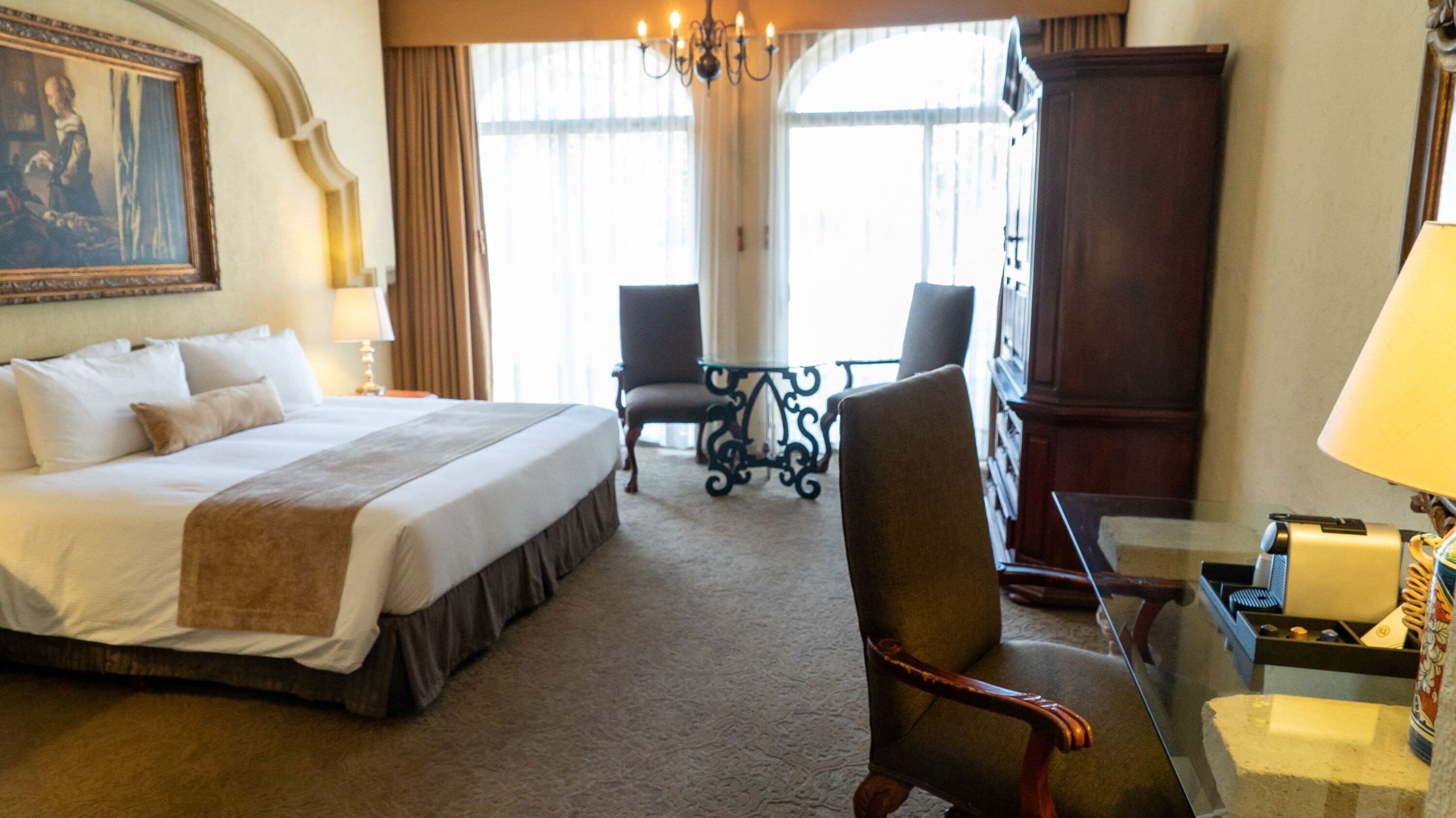 Une chambre d’hôtel avec un grand lit, deux fauteuils, une petite table, un lustre et un tableau au mur.
