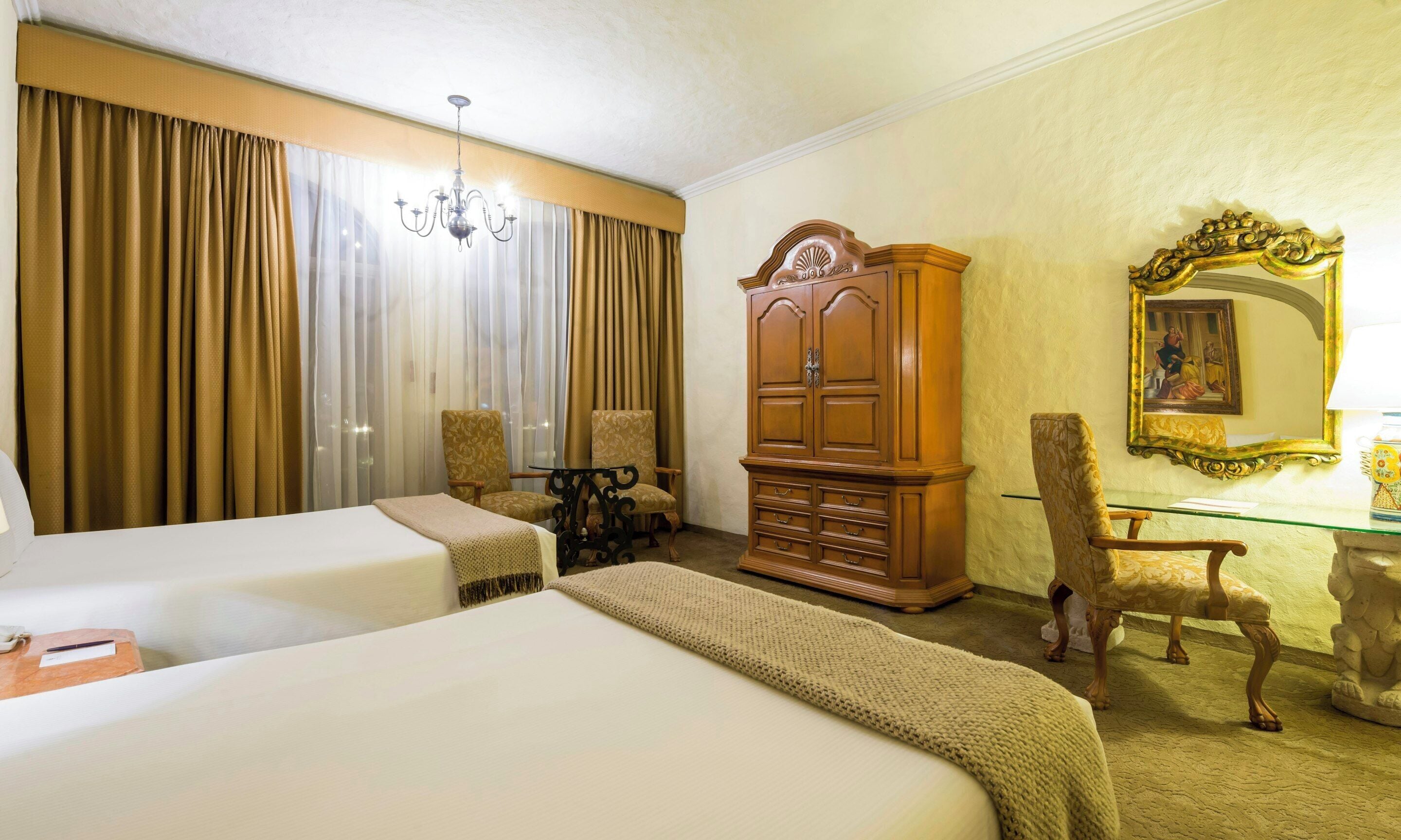 A hotel room with two beds, a wooden wardrobe, a chair, a glass table, and a mirror with a painting.
