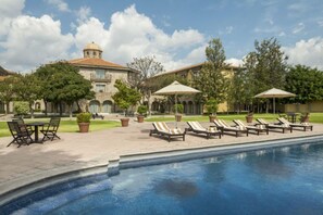 A pool area with lounge chairs and umbrellas, surrounded by a courtyard with trees and a building with a dome.