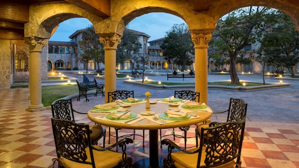 Un área de comedor al aire libre con una mesa redonda dispuesta para seis personas, rodeada de sillas ornamentadas, bajo un arco de piedra con vista a un patio y edificios.