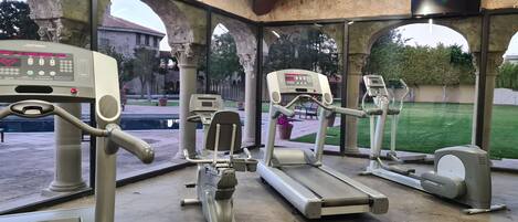Uma academia ao ar livre com equipamentos de ginástica, incluindo esteiras e um remo, sob uma área coberta com colunas de pedra e vista para um pátio.