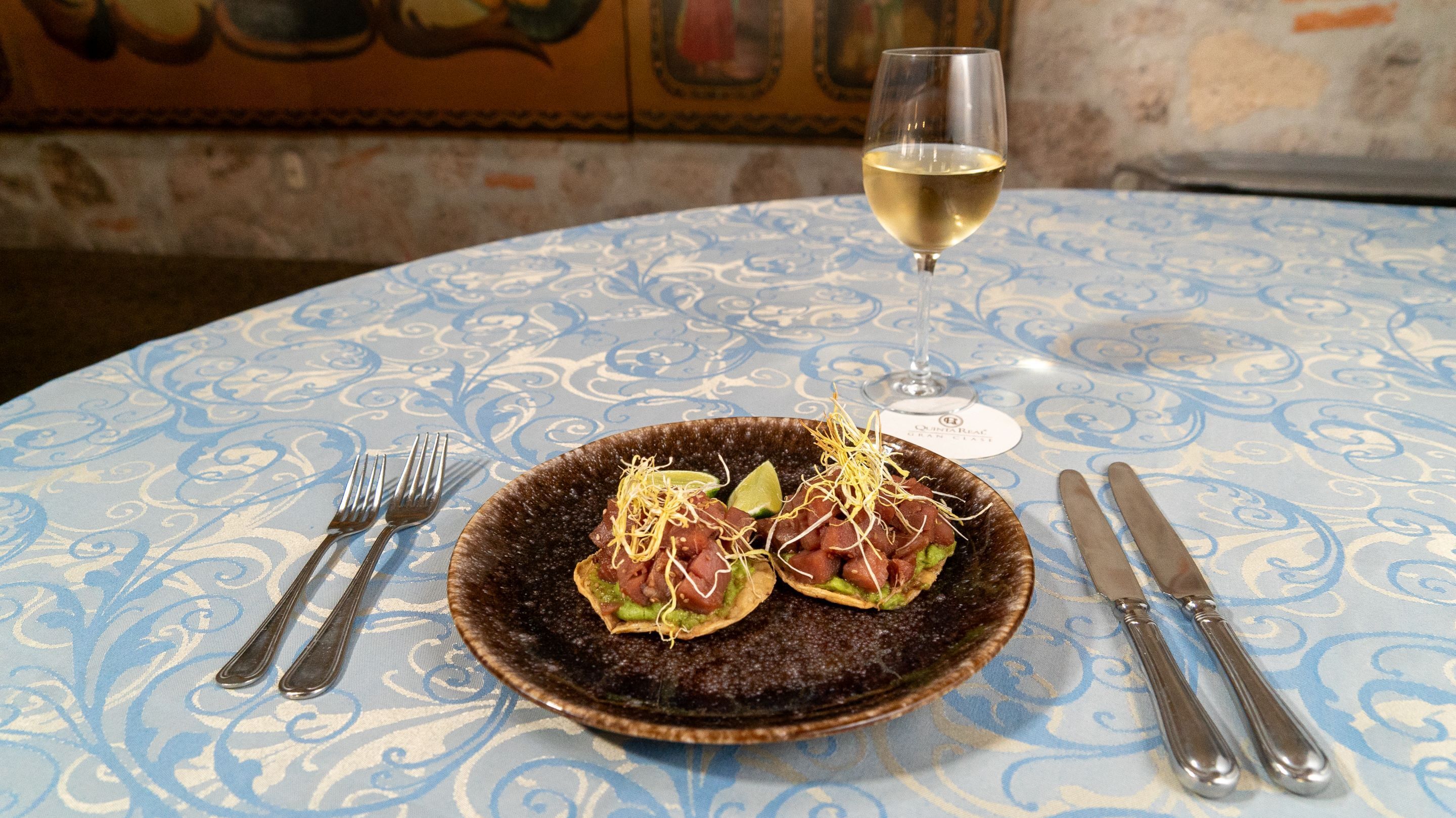 Een bord met twee taco's op een geruit tafelkleed, met een glas witte wijn, bestek en een menukaart.