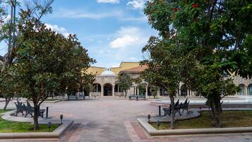 Un patio con un espacio abierto central, rodeado de bancos y árboles, que conduce a un edificio con arcos y techo de tejas.