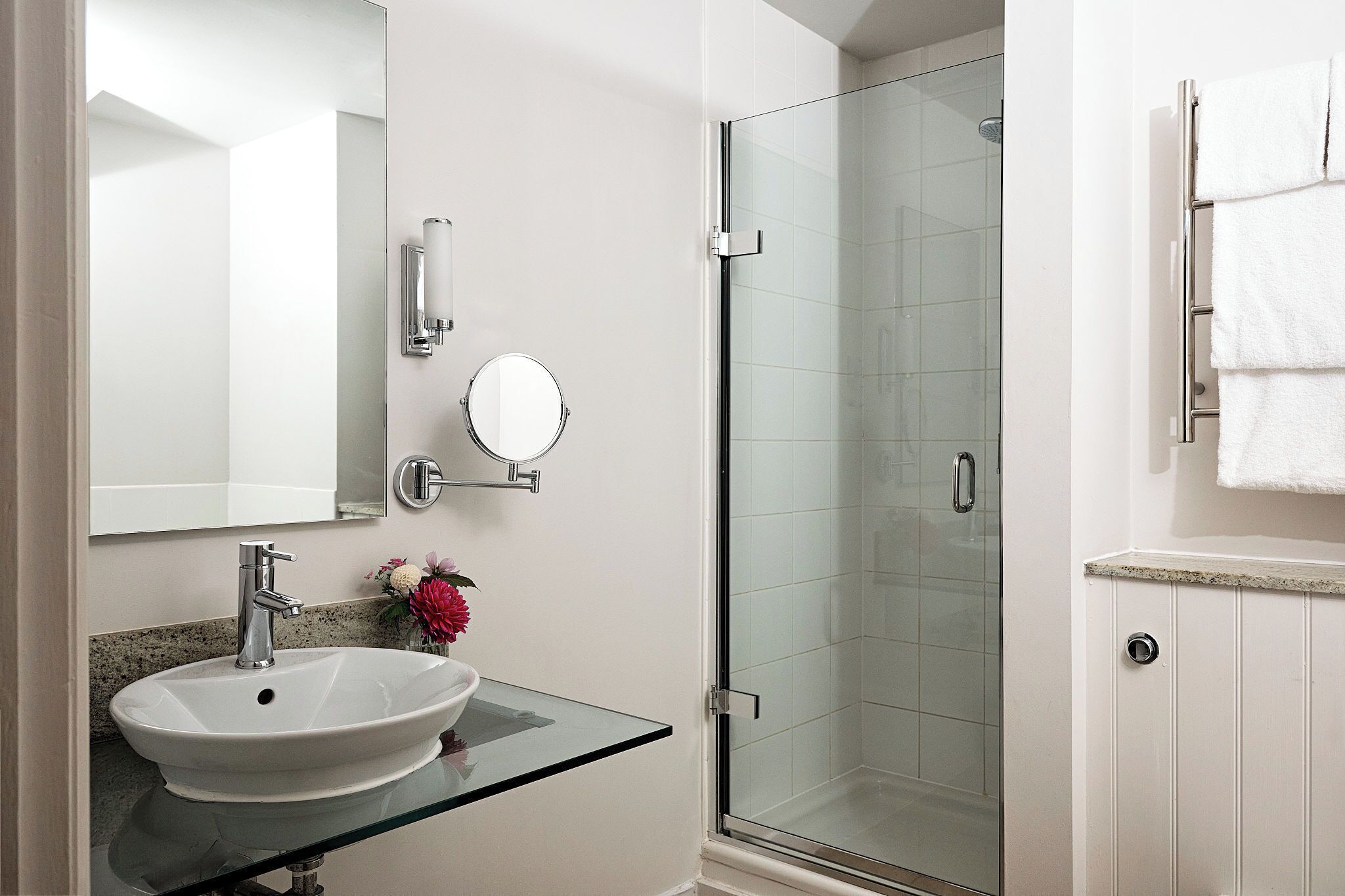 A bathroom with a glass-enclosed shower, a white sink with a glass countertop, a round mirror, and a towel rack.