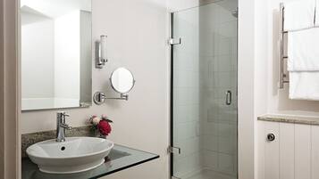 A bathroom with a glass-enclosed shower, a white sink with a glass countertop, a round mirror, and a towel rack.