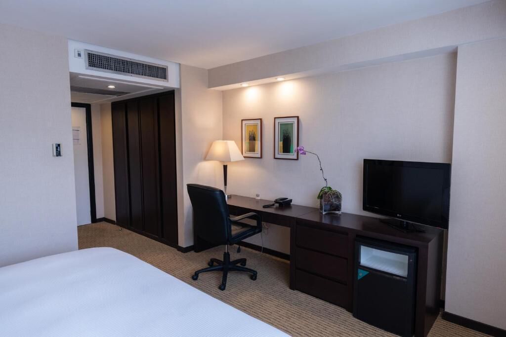A hotel room with a bed, a desk with a chair, a television, a lamp, and two framed pictures on the wall.