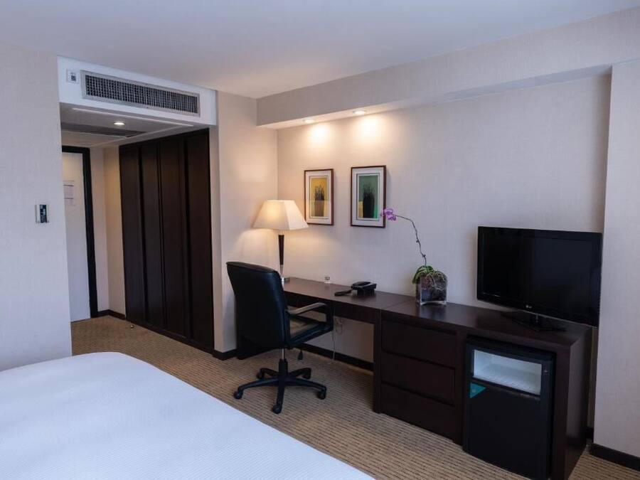 A hotel room with a bed, a desk with a chair, a television, a lamp, and two framed pictures on the wall.