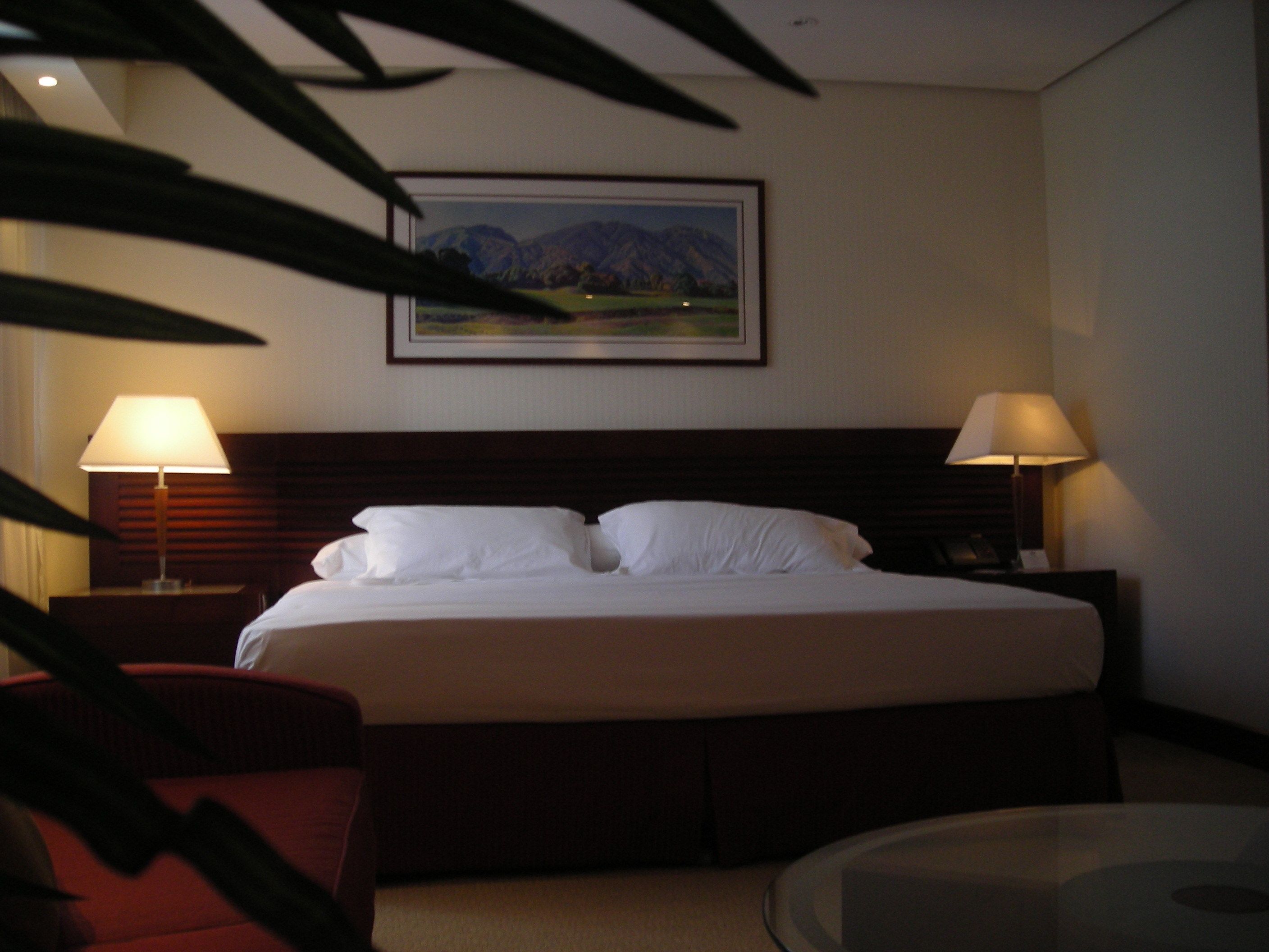 Ein Hotelzimmer mit einem großen Bett, zwei Nachttischlampen, einem gerahmten Landschaftsbild und einem Glastisch.