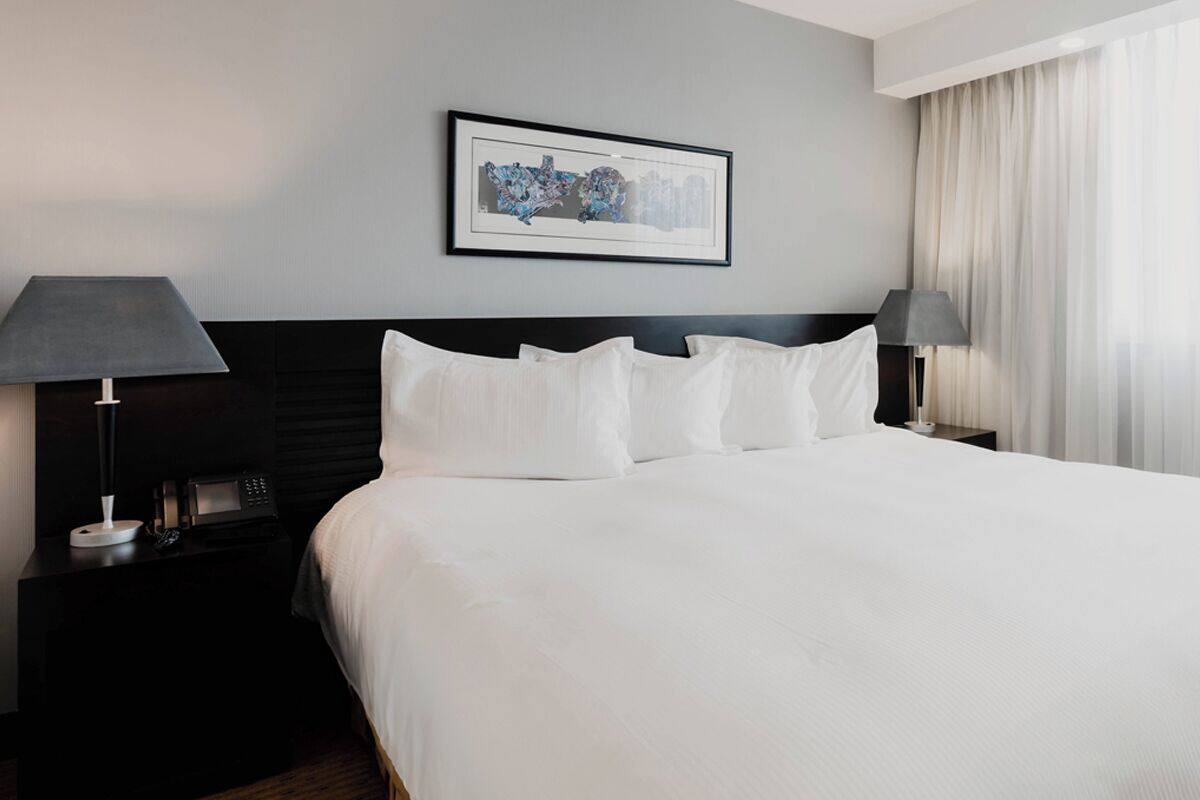 Une chambre d’hôtel avec un grand lit, une tête de lit sombre, une œuvre d’art encadrée au mur et une table de chevet avec une lampe et un téléphone.