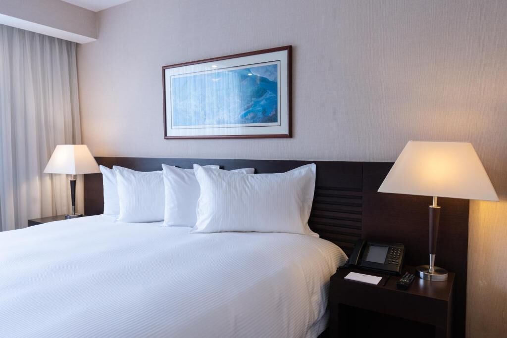 Een hotelkamer met een groot bed, twee bedlampjes, een kunstwerk in een lijst aan de muur, en een nachtkastje met een telefoon en notitieboekje.