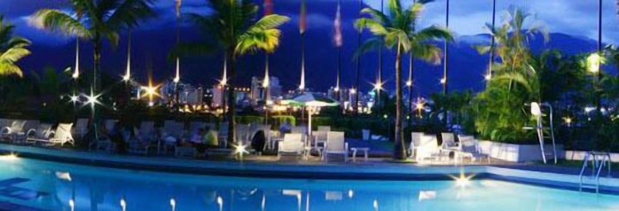 A swimming pool with lounge chairs and palm trees, surrounded by a resort area with buildings and a night sky.