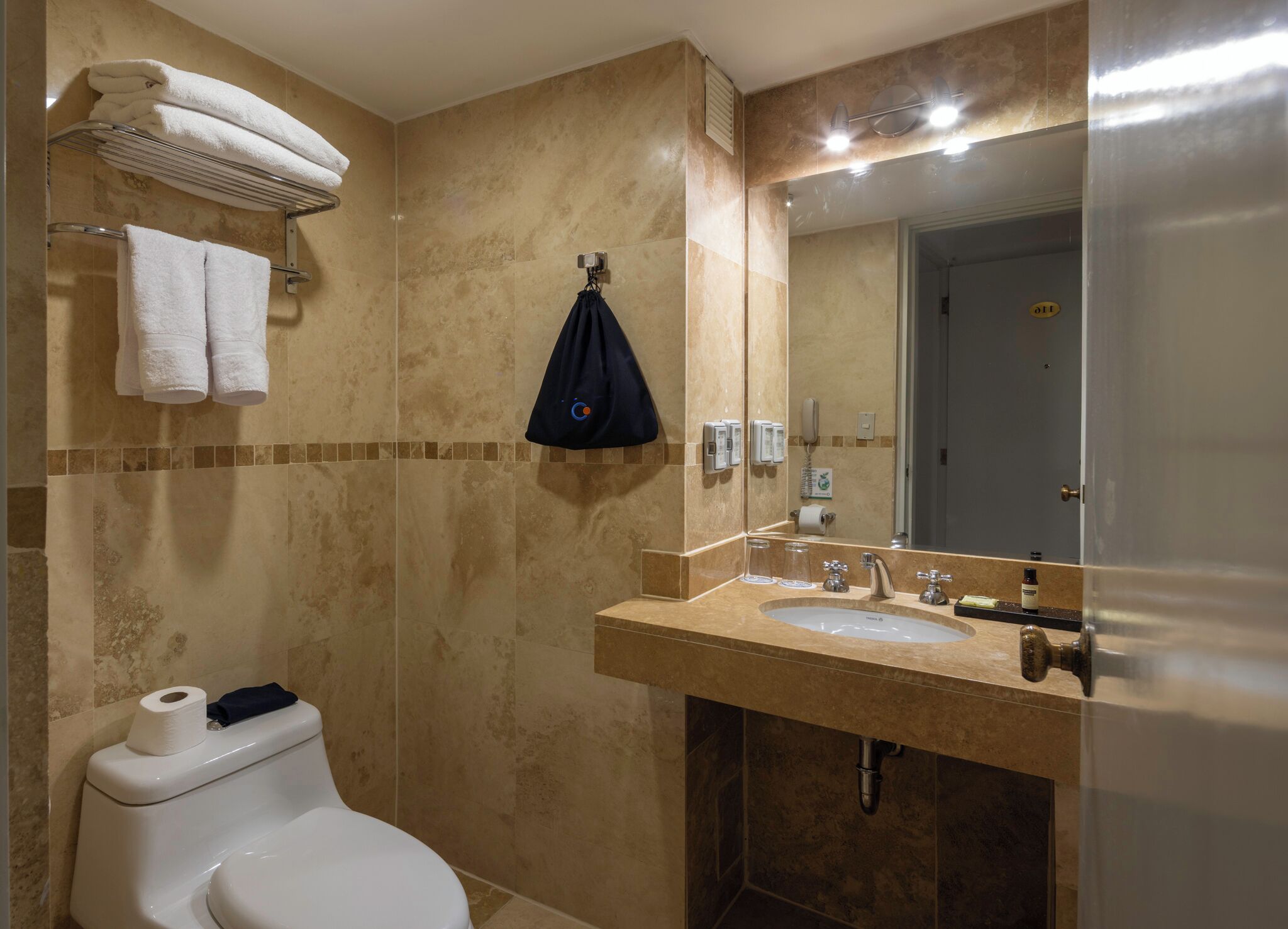 A bathroom with a toilet, sink, and mirror. There are towels on a rack and a black bag hanging on the wall.
