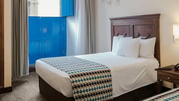 Habitación de hotel con una cama grande, cabecero de madera, araña y vista a un edificio azul a través de una ventana.