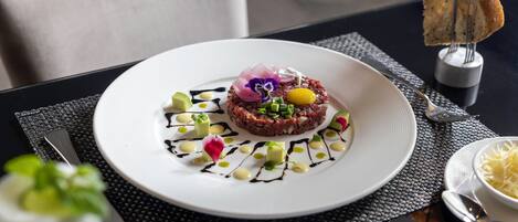 Un plato de tártara de carne con adornos y salsas sobre un plato blanco, acompañado de un tenedor y un cuchillo sobre una mesa negra.